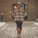 a woman wearing a plaid hoodie, beanie and tall boots walking down a brick street