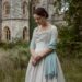 a woman in a white Regency dress with a light blue shawl in front of a castle