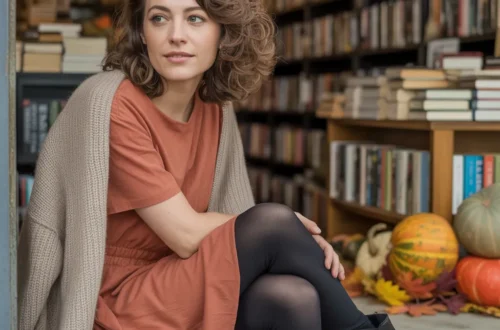 A soft-focus portrait photograph of a woman, aged 25 to 30, with medium-length, gently wavy brown hair framing her face as she gazes slightly off-camera with a thoughtful expression. She is wearing a casual rust-terracotta dress with short sleeves, layered with an open-front beige cardigan, black leggings, and sleek black Chelsea boots, conveying a relaxed yet stylish aesthetic. Behind her, a warmly lit bookstore displays shelves overflowing with books and autumnal decorations like pumpkins and scattered leaves, suggesting a cozy, inviting atmosphere.