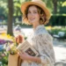 A woman wearing a white floral dress, straw hat holding a woven tote and a book in a sunny park.