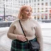 close up of a 25-year-old plus size white woman with soft blonde hair smiles gently, looking off to one side. She wears a cream ribbed turtleneck sweater tucked into a navy and green plaid wool skirt, thick black tights, black ankle boots, and a small black crossbody bag. She accessorizes with pearl stud earrings. The background is a brightly lit snowy city plaza decorated with string lights.