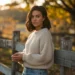 a woman in a sweater and jeans leaning against a wooden fence in the fall