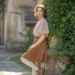 Woman in blush pink puffy-sleeve blouse and brown corduroy midi skirt with cream tights and ankle boots standing against ivy-covered stone wall, natural no-makeup look, cottagecore style.
